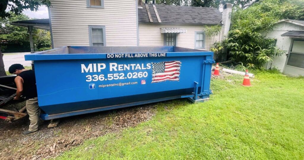 A blue dumpster with an American flag design on the side, placed in front of a home undergoing renovation. The sign above the dumpster reads 'Leave This Line.' The area around the dumpster is filled with construction debris and materials, making it an ideal choice for an affordable dumpster rental for home renovation in Reidsville.