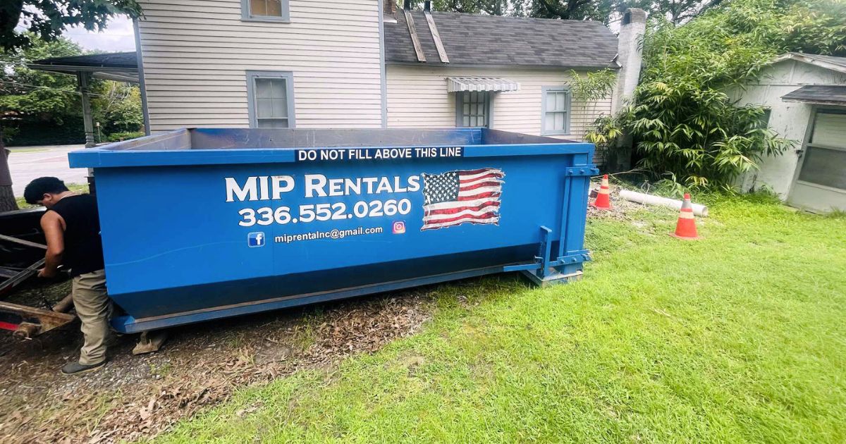 A blue dumpster with an American flag design on the side, placed in front of a home undergoing renovation. The sign above the dumpster reads 'Leave This Line.' The area around the dumpster is filled with construction debris and materials, making it an ideal choice for an affordable dumpster rental for home renovation in Reidsville.
