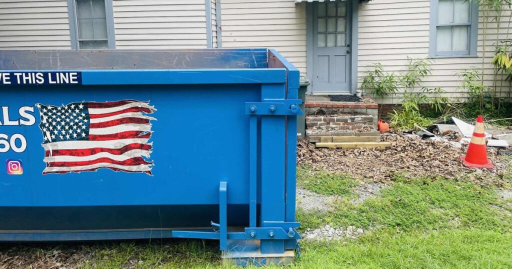 Blue dumpster with an American flag design on the side, placed outside a home under renovation. The sign above the dumpster reads 'Leave This Line.' Nearby, there are construction materials, including bricks and wooden boards, scattered on the ground. This affordable dumpster rental is ideal for home renovation projects in Reidsville.